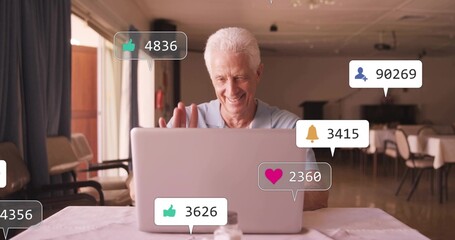 Waving senior man in light-blue polo using silver laptop in dining area, notifications, salt shaker