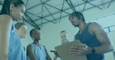 Talking coach in dark tank holding clipboard at gym with players in light-blue jerseys, copy space