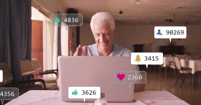 Waving senior man in light-blue polo using silver laptop in dining area, notifications, salt shaker - Powered by Adobe