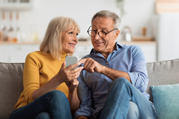 A smiling couple sits closely on a couch, sharing a lighthearted moment as they look at a smartphone together in a warm and inviting living room filled with natural light.