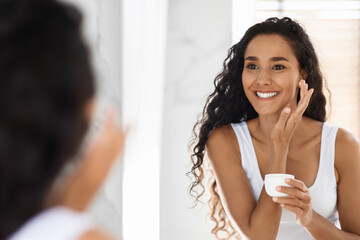 A woman smiles at her reflection while applying cream to her face in a well-lit bathroom. She has...