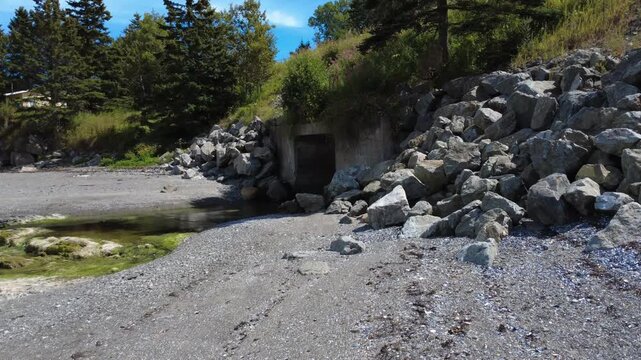 A coastal landscape with the outlet of a concrete culvert surrounded by large rocks. A calm stream, the pebbly shoreline and vegetation in the background.