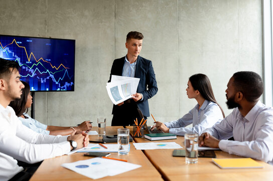 In a modern office, a disappointed boss shares financial reports with a group of diverse coworkers. They brainstorm solutions to address the challenges reflected in the data displayed.