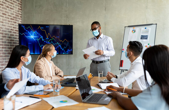 A group of diverse business professionals is holding a meeting in a modern office. A Black businessman in a medical mask presents strategies while colleagues listen attentively.