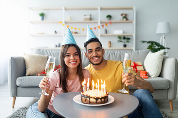 A young couple enjoys a joyful birthday celebration in a bright living room. They hold champagne glasses and smile at the camera while surrounded by gifts and a cake with candles.