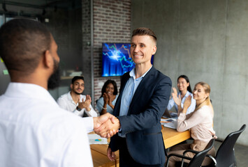 A male HR manager shakes hands with an African American employee in an office setting. Colleagues applaud as they welcome the new team member during a corporate meeting in the boardroom.