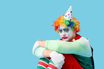 Young man dressed as sad clown sitting on blue background. April Fool's day celebration