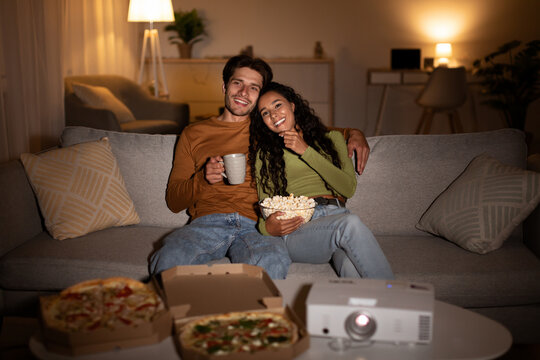 A couple sits on their couch enjoying a movie night at home. They smile while holding coffee and popcorn, surrounded by pizza boxes and a projector, creating a cozy atmosphere together.