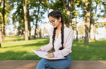 Obraz premium A young woman is seated on a wooden platform in a park, focused on writing in her notebook. She enjoys the warm sunlight and greenery around her.