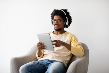 A cheerful young Black man sits comfortably in a gray armchair, enjoying his digital tablet while wearing wireless headphones. He is browsing the internet and smiling.