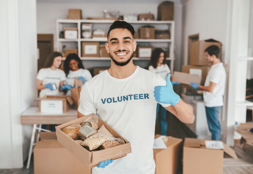 A group of volunteers works in a community center, sorting food donations. One volunteer holds a box of food, smiling and giving a thumbs up, showing support and teamwork.