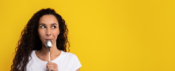 A woman with curly hair stands in front of a bright yellow background holding a spoon to her lips...