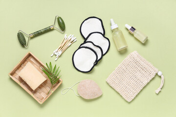 Reusable cotton pads with bottles of cosmetic products, soap bar and face massage roller on green background