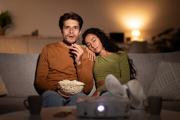Husband watches a movie using a projector in their living room at night. His wife sleeps beside him...