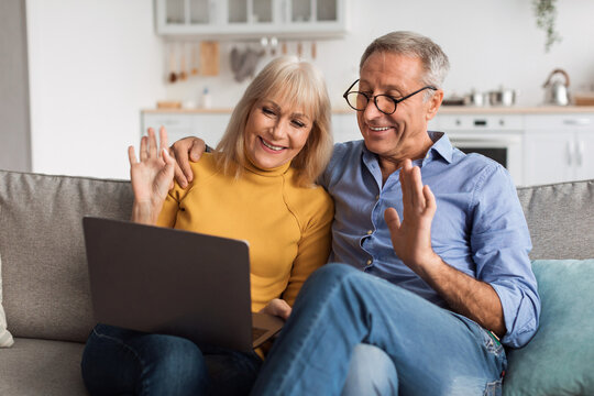 A happy couple enjoys a video call from their comfortable couch in a bright living room. They smile and wave at the screen, sharing a warm moment together.