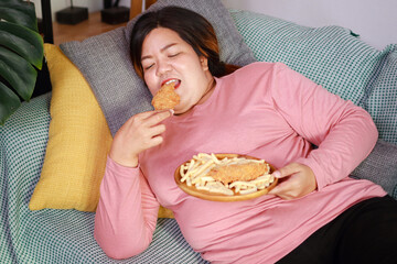 Fat Asian woman lying on the couch eating fried chicken and french fries. Fast food, junk food. Obesity concept.