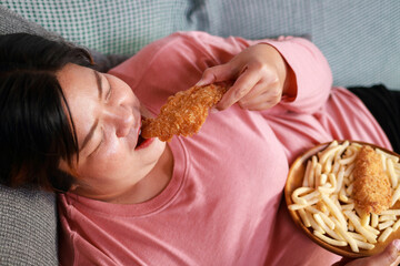 Fat Asian woman lying on the couch eating fried chicken and french fries. Fast food, junk food. Obesity concept.