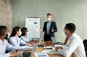 Businessmen and women in masks gather in a modern office to discuss company strategy. A young CEO leads the meeting, presenting ideas to diverse coworkers.