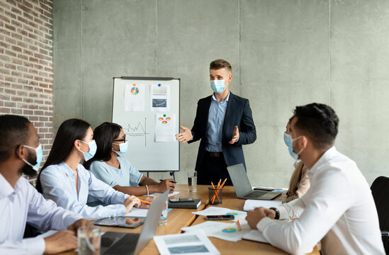 Businessmen and women in masks gather in a modern office to discuss company strategy. A young CEO leads the meeting, presenting ideas to diverse coworkers.