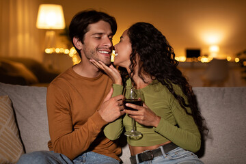 Spouses share a warm moment at home, sipping red wine and enjoying each other's company in a cozy living room at night. The scene captures intimacy and romance.