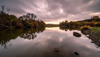 A tranquil lake scene at sunset, with reflections of trees and a dramatic, cloudy sky.