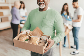 A man holds a box filled with food items while smiling. In the background, others are preparing packages for distribution in a community center focused on helping those in need.