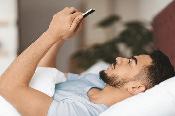 Fototapeta premium A young man with a beard relaxes in bed while checking his smartphone. Soft morning light filters into the room, creating a calm and cozy atmosphere as he scrolls through his device.