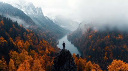 A man stands on a mountain peak, overlooking a valley filled with autumn foliage and a river, shrouded in fog.