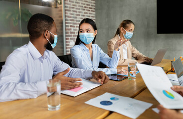 Business people in protective face masks engage in discussion at a corporate meeting. The group, representing various backgrounds, works together in an office setting amid the pandemic.
