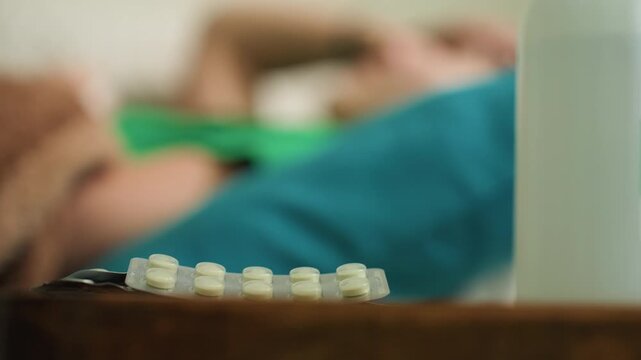 medicine setup indoors, focus on pills with woman blurred behind, scene depicting medication arrangement on table at home setting, image shows medicine pills on table with woman relaxing in background