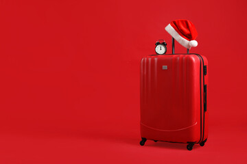 Christmas vacation. Suitcase with Santa hat and alarm clock on red background, space for text