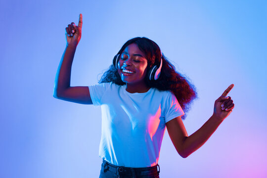A young woman enjoys dancing while wearing headphones, immersed in vibrant blue and pink lighting. Her joyful expression reflects love for music in an energetic atmosphere.