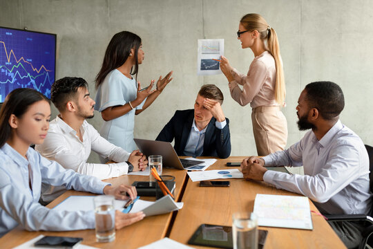 Two female employees are arguing during a corporate meeting as a multiethnic group of colleagues observes. Tension arises over a new project in a boardroom filled with documents and technology.