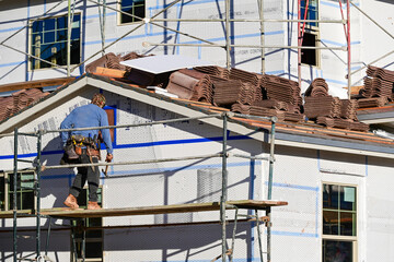 Construction worker standing on scaffolding beside a house with stacked roof tiles, illustrating residential building activity, exterior preparation, and roofing installation in a new home project © Eduardo Barraza