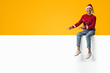 A cheerful man wearing a Christmas sweater and Santa hat sits on a white wall. He is pointing and smiling, surrounded by a bright yellow background that adds festive cheer.