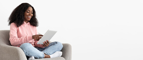 A joyful young woman sits cross-legged on a couch, wearing a pink hoodie and jeans, engaging with her tablet in a well-lit, minimalist space, smiling with delight.