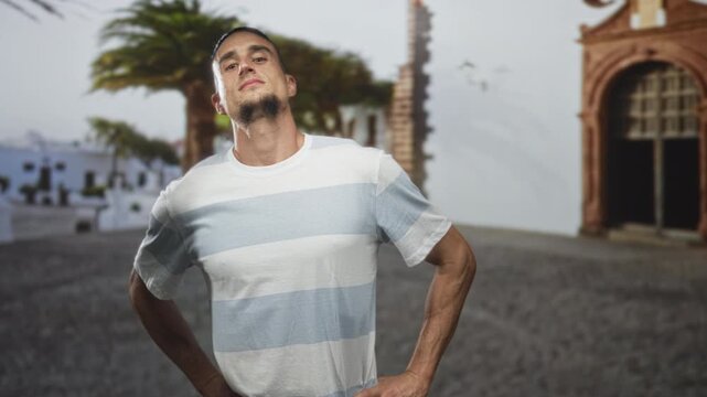 Man hands on hips posing in a sunlit street plaza beside an arched building, striped tshirt visible; confidence poise.