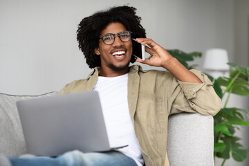 Smiling black man engages in a phone conversation while working on his laptop at home. He appears relaxed on his couch in a bright living room, showcasing the comfort of remote work.