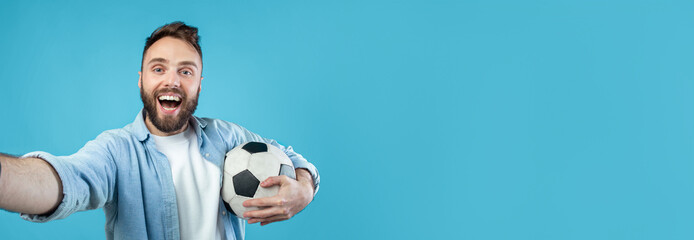 A young man with a beard enjoys holding a soccer ball and taking a selfie. His excitement is evident as he stands against a vibrant blue backdrop, suggesting a fun moment during a casual day.