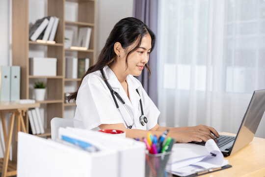 Asian Young female medical professional with stethoscope working on laptop at bright desk, focused on patient notes and digital records in modern home office setting