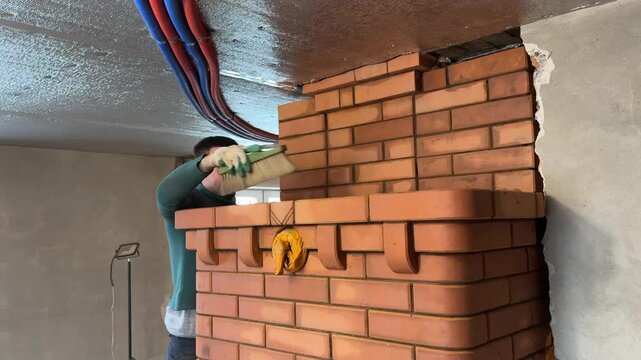 A craftsman cleans the joints of fresh brickwork using a metal brush. Construction of a heating stove.