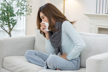 Asian woman sneezing into tissue while sitting on sofa, showing symptoms of cold, flu, or seasonal allergy infection at home, Health care and allergy concept
