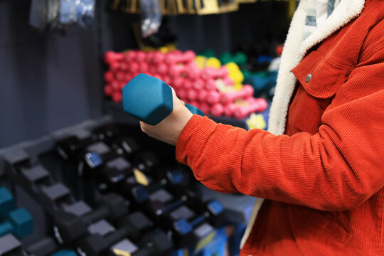 Man in a red jacket choosing a blue dumbbell in a sports equipment store with colorful weights in the background.
