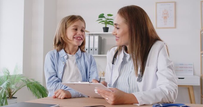 Friendly female pediatrician sitting at desk with smiling child patient, listening to complaints during examination in medical clinic. Cheerful boy talking to kind doctor about his health problems.