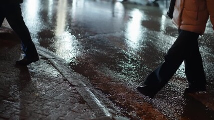 Pedestrians stride along a rain-soaked city sidewalk at night, legs crossing glistening pavement as blurred headlights and taillights shimmer across wet asphalt in urban downpour - Powered by Adobe