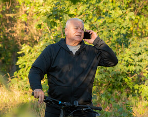 An elderly gray-haired man uses a smartphone while cycling through the forest.