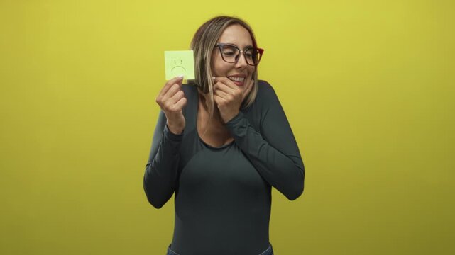 Woman holding a sad sticky note while biting her nails and wearing glasses in studio with yellow backdrop; anxiety vulnerability.