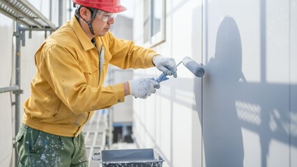Construction worker in yellow jacket and red hard hat painting wall with roller protective gear visible asian asia chinese