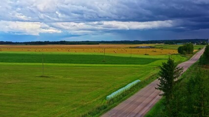 Country road with hay bales and darkening storm clouds over flat farmland in aerial pedestal up