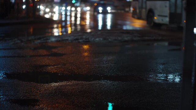 Rain-soaked city street at night with deep potholes and shimmering reflections from headlights and streetlights, as cars stream through a slick intersection.